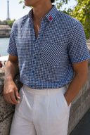 A man wearing a blue and white gingham short-sleeve button-up shirt with a chest pocket and white trousers, leaning against a stone wall outdoors near water and greenery.
