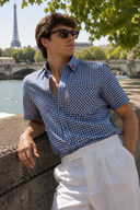 A man wearing a blue and white gingham short-sleeve button-up shirt and white trousers leans against a stone wall outdoors in Paris, with the Eiffel Tower and water in the background.