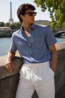 A man wearing a blue and white gingham short-sleeve button-up shirt and white trousers leans against a stone wall outdoors, with the Eiffel Tower and water in the background.