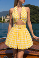 A woman wearing a yellow and white gingham two-piece swimsuit with a halter top and high-waisted flared skirt, standing on a wooden boat with water and a scenic lake background.