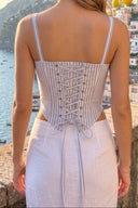 A woman stands outdoors by a stone wall overlooking a coastal town and sea, shown from the back wearing a light-colored sleeveless dress with thin straps, a striped corset-style back panel with lace-up detailing, and a pale skirt. Lemon branches hang overhead.