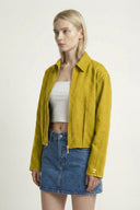 Woman wearing a mustard yellow jacket over a white top and blue denim skirt on a plain background