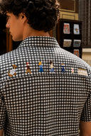 A man viewed from behind wearing a black and white gingham short-sleeve button-up shirt with embroidered cartoon characters across the upper back.