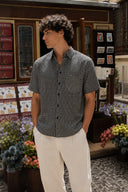 A man wearing a black and white micro-patterned short-sleeve button-up shirt and off-white trousers, standing in front of a shop with flowers.