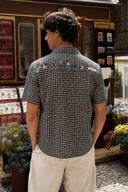 A man viewed from behind wearing a black and white micro-patterned short-sleeve button-down shirt with colorful embroidered figures across the upper back, paired with cream-colored pants, standing in front of a shop counter.