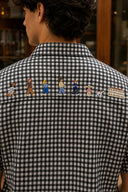 A man viewed from behind wearing a black and white gingham short-sleeve shirt with a row of colorful embroidered figures and a small building motif across the upper back.