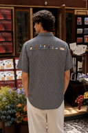 A man viewed from behind wearing a black and white gingham short-sleeve button-up shirt with a row of colorful pins across the upper back, paired with off-white trousers, standing in front of a shop display.