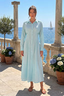 A woman stands on a sunlit terrace overlooking the blue sea, wearing a light blue and white gingham long-sleeve midi shirt dress with a collar and button front.