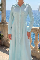 A woman wearing a light blue and white gingham long-sleeve shirt dress, standing on a stone balcony with the blue sea and a sailboat in the background.