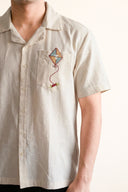 Beige shirt with embroidered kite on the chest pocket against a white background