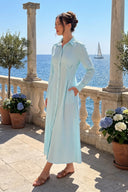 A woman wearing a light blue and white gingham long-sleeve midi shirt dress stands on a stone terrace overlooking the blue sea, with columns, potted plants, and a sailboat visible in the background.
