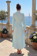 A woman stands on a sunlit terrace with a sea view behind her, wearing a light blue and white gingham long-sleeve midi dress with a wrap-style back and belt. Stone columns, potted flowers, and a sailboat are visible in the background.