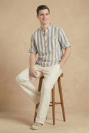 Man sitting on a wooden stool wearing a striped shirt and beige pants against a beige background